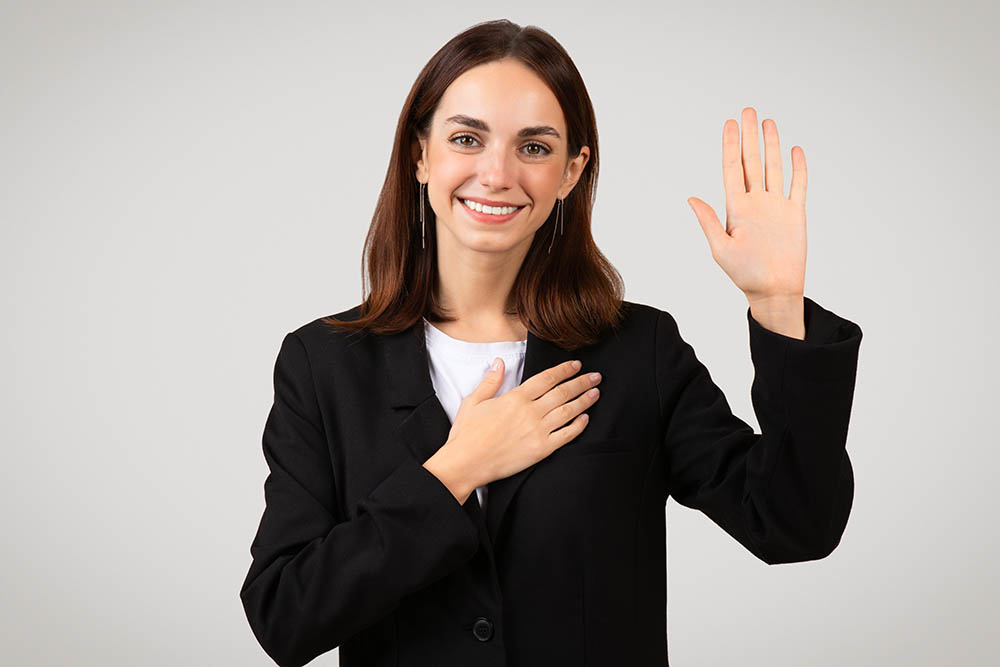 Pleasant millennial caucasian businesswoman with a sincere smile raises her right hand to take an oath or pledge, placing her left hand over her heart, signifying honesty and commitment