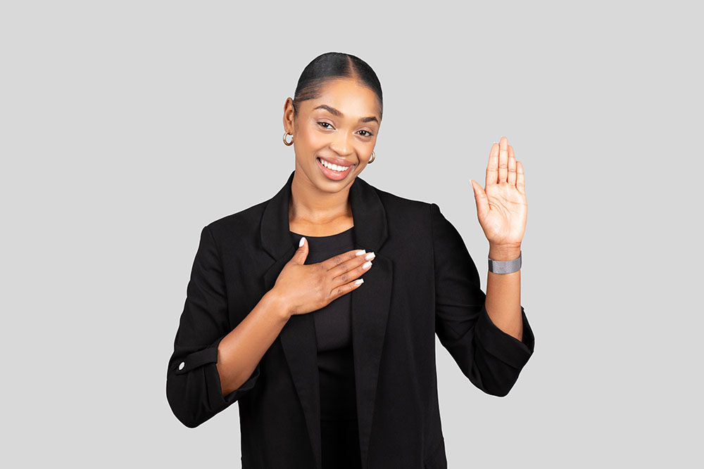 Radiant young woman in black business attire smilingly takes an oath with her right hand raised, emanating trustworthiness and professionalism, isolated on gray studio background