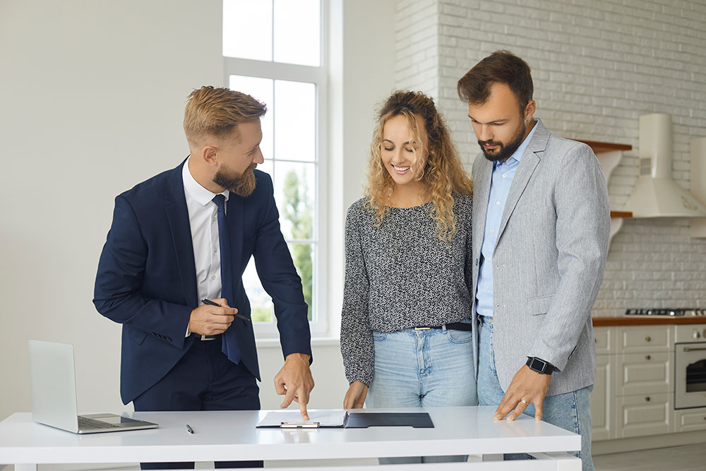 Young married couple buying or renting house. Husband and wife standing at table in kitchen in their new apartment and signing contract given by realtor or real estate agent